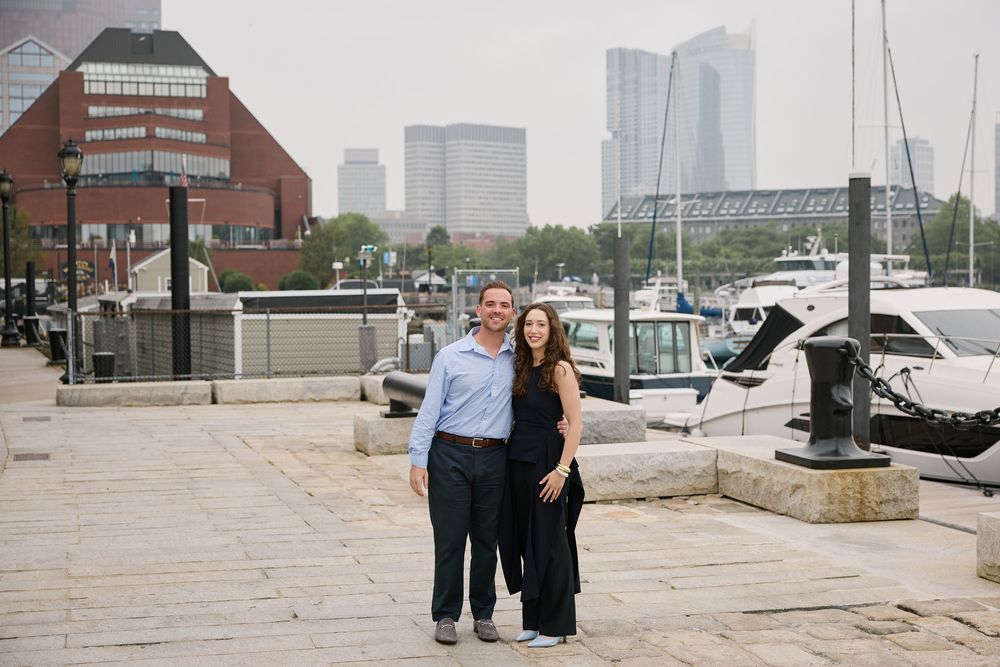 Couple smiling together on a waterfront promenade with marina boats and city skyline in the background.