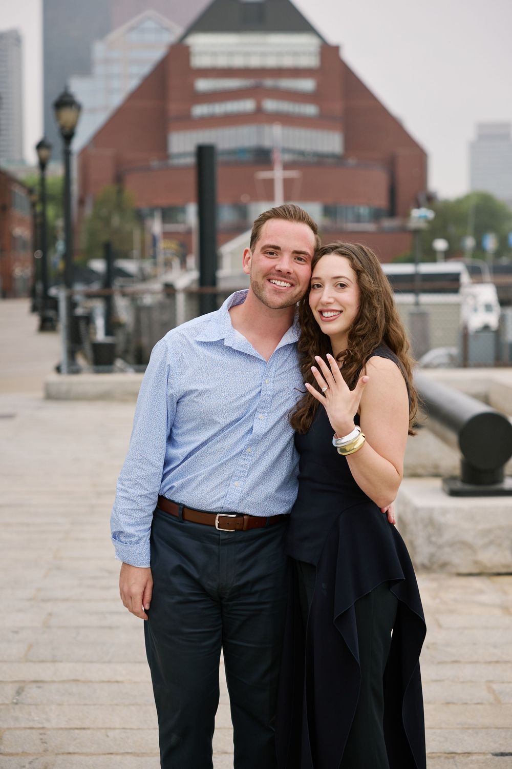 Happy couple showing off engagement ring on city waterfront steps with urban skyline backdrop.