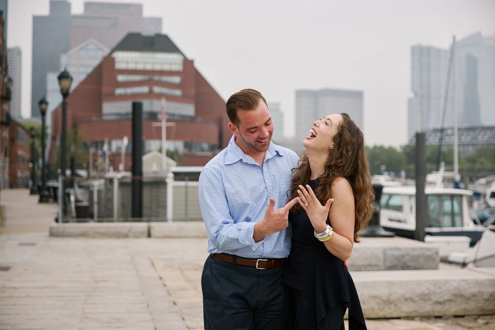 Joyful couple laughing together at harbor waterfront during engagement photo session.