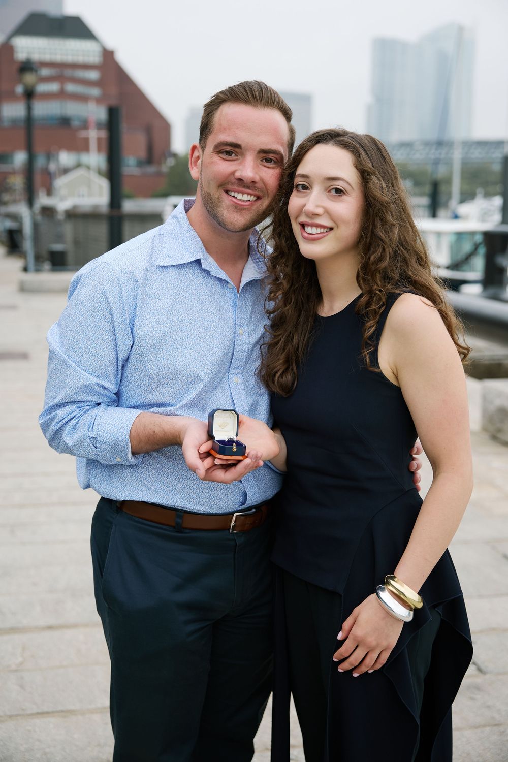 Smiling couple posing outdoors holding an engagement ring box near a waterfront with city buildings in the background.