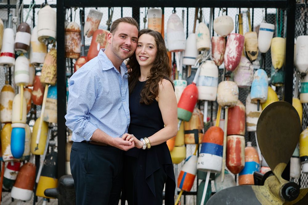 Couple smiling and holding hands in front of colorful nautical buoys display at a coastal location.