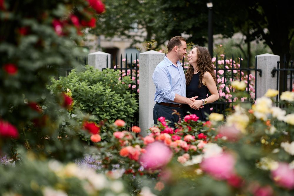 Couple sharing a romantic moment surrounded by colorful roses in a lush garden with white fence posts.