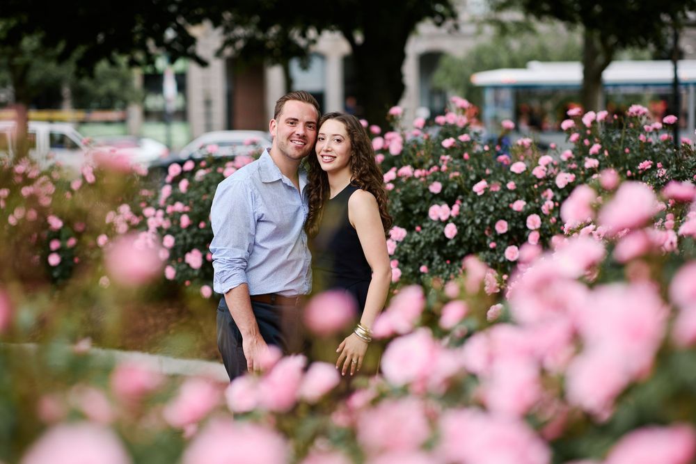 Smiling engaged couple posing romantically among blooming pink roses in garden park.
