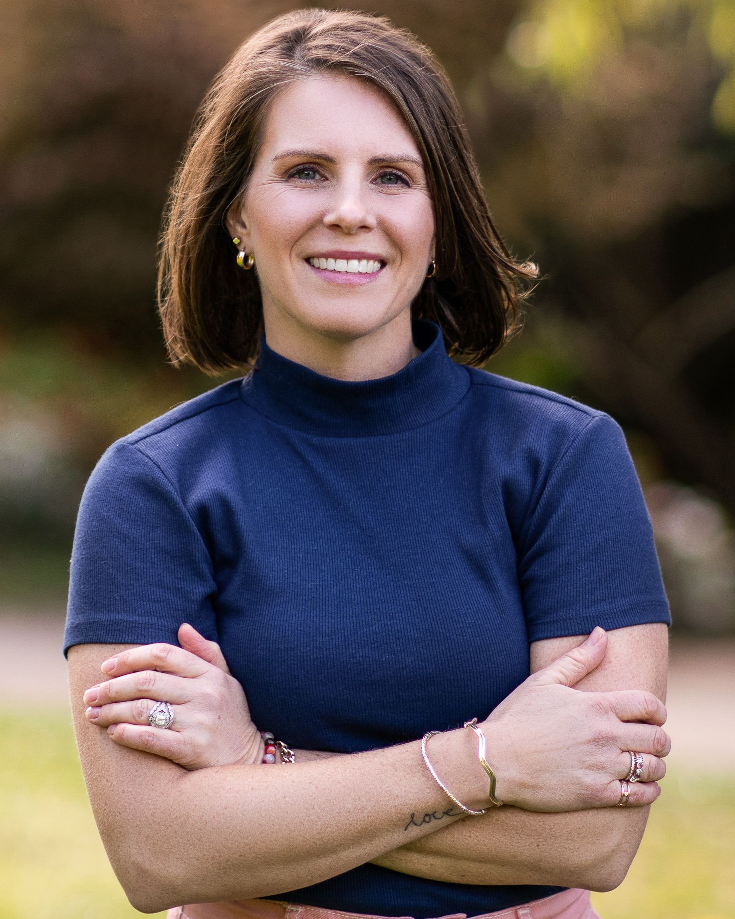 Professional woman with brown hair smiling confidently, arms crossed, wearing navy top outdoors.