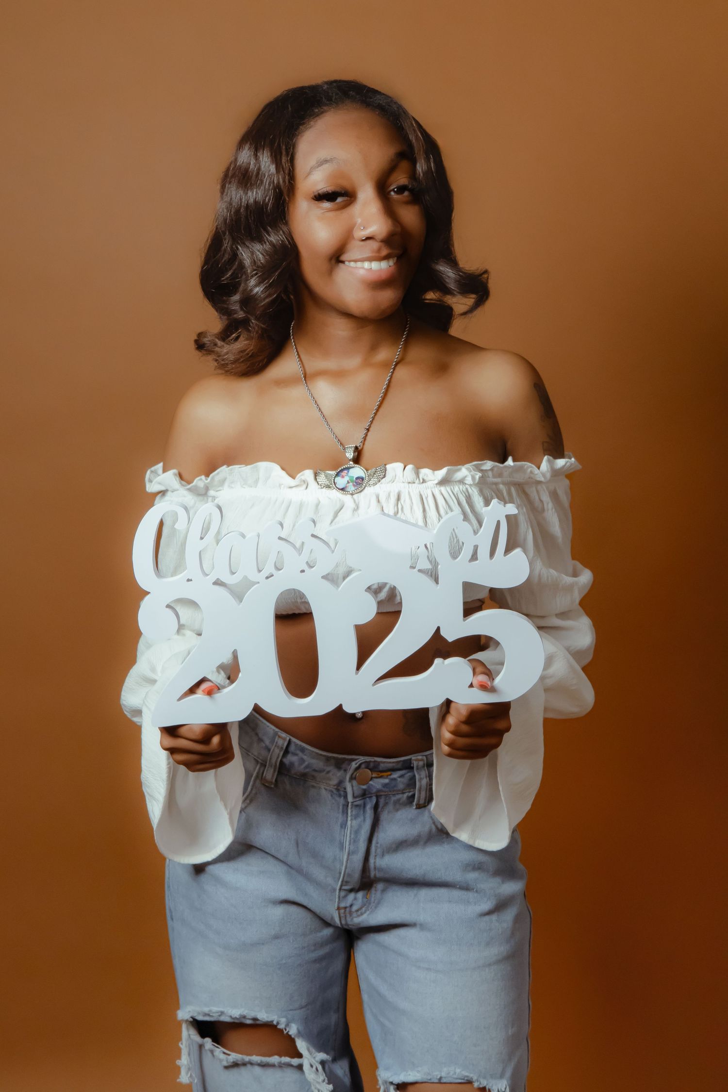 Smiling young woman holding a white Class of 2025 sign, wearing off-shoulder top and ripped jeans on brown background.