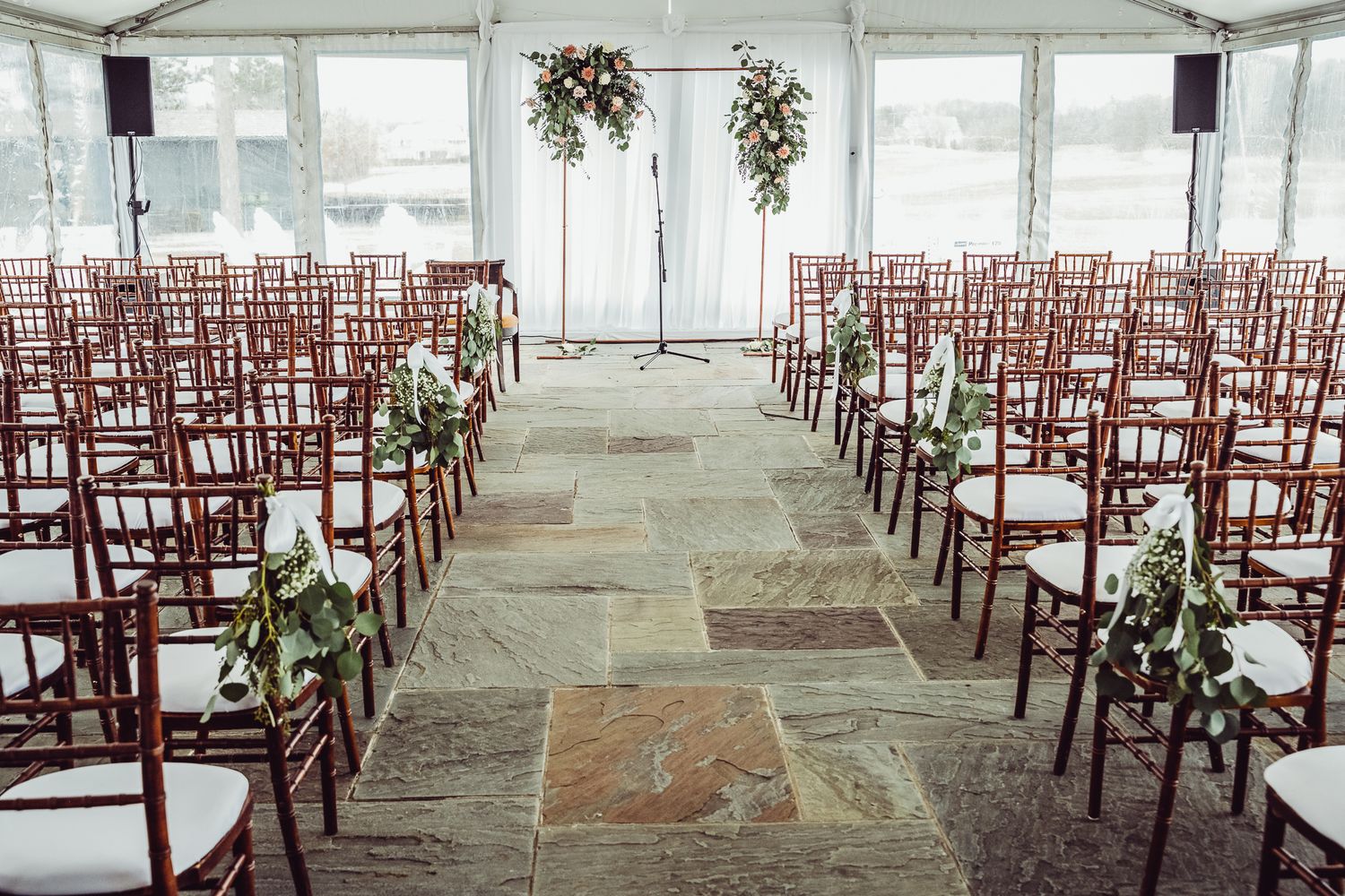 Elegant wedding ceremony setup under a white tent with mahogany Chiavari chairs, stone aisle, and floral arch backdrop.