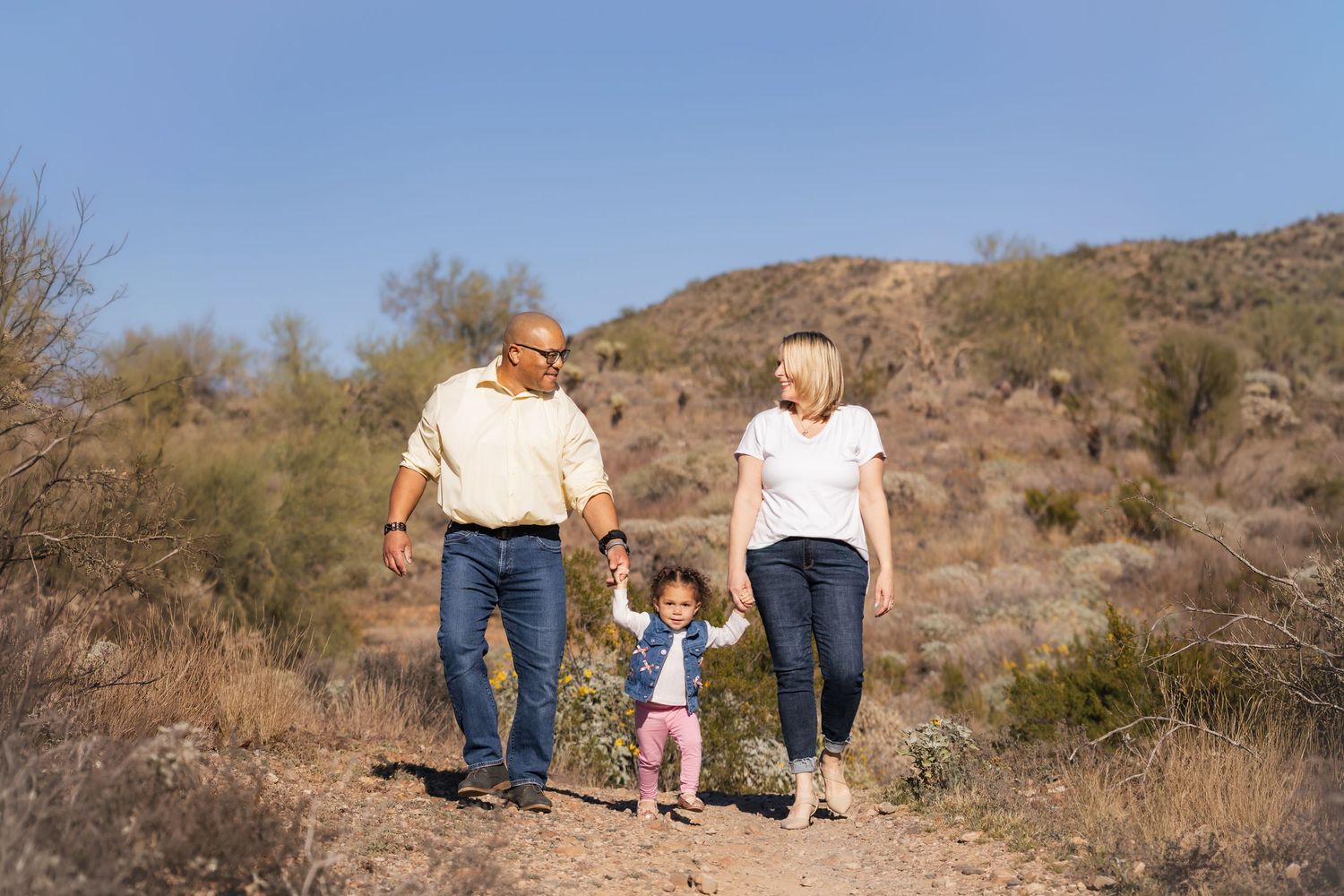 Family of three hiking on a desert trail, grandparents holding hands with toddler girl against blue sky and hills.