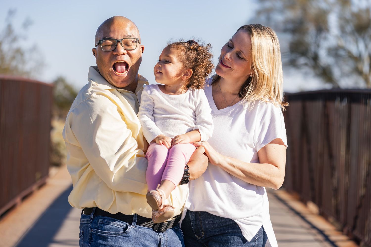 A joyful interracial family with a man, woman, and curly-haired toddler laughing together on a sunny bridge outdoors.