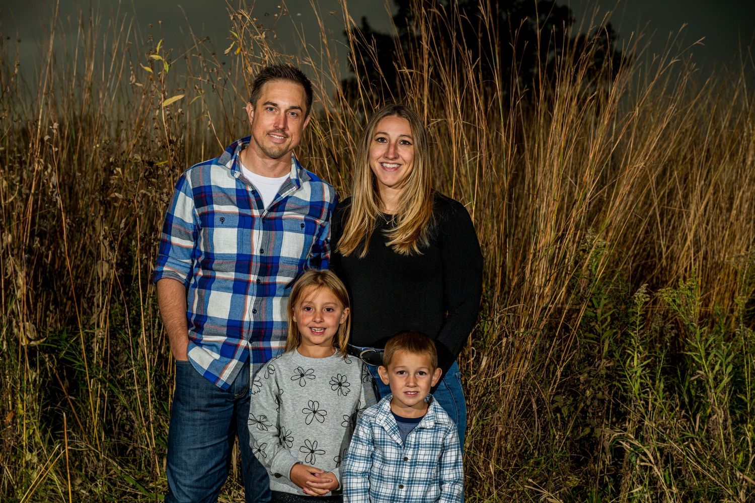 Family of four posing outdoors near tall golden grasses, two adults and two children smiling for a portrait photo.