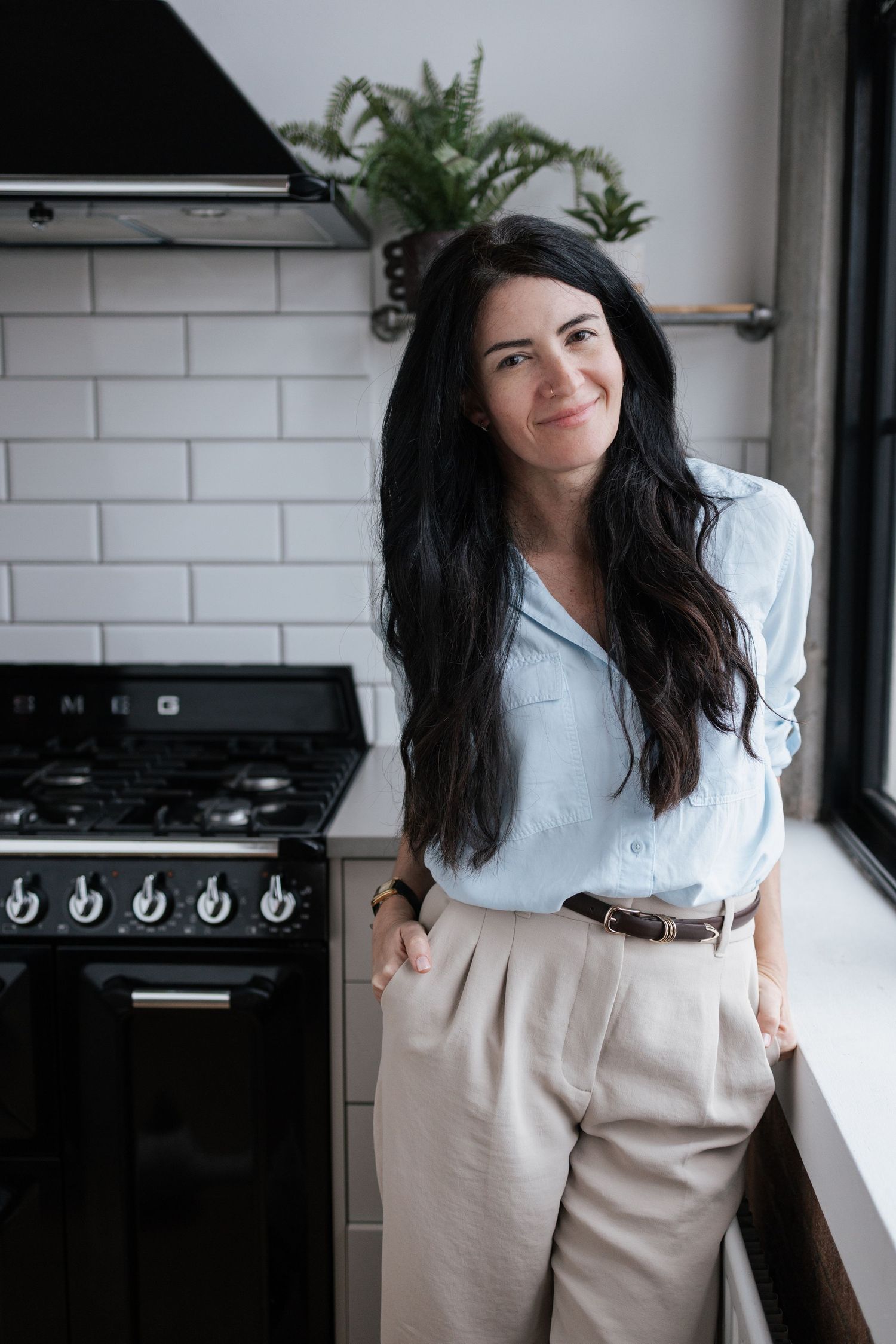 A dark-haired woman in a light blue shirt and beige trousers stands smiling in a modern kitchen with a black stove.