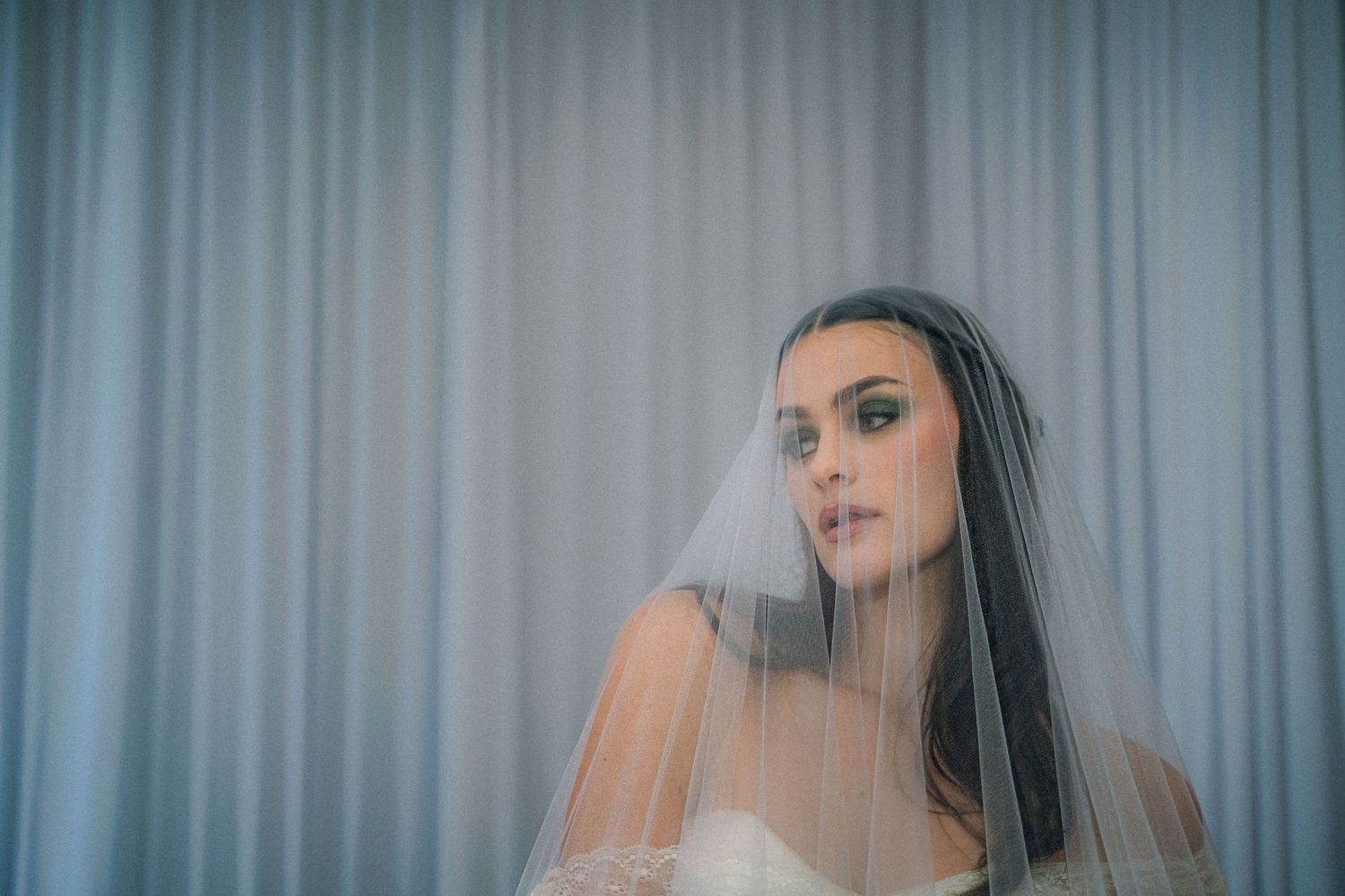 Bride wearing a sheer white veil and strapless gown poses elegantly against a soft gray draped curtain backdrop.