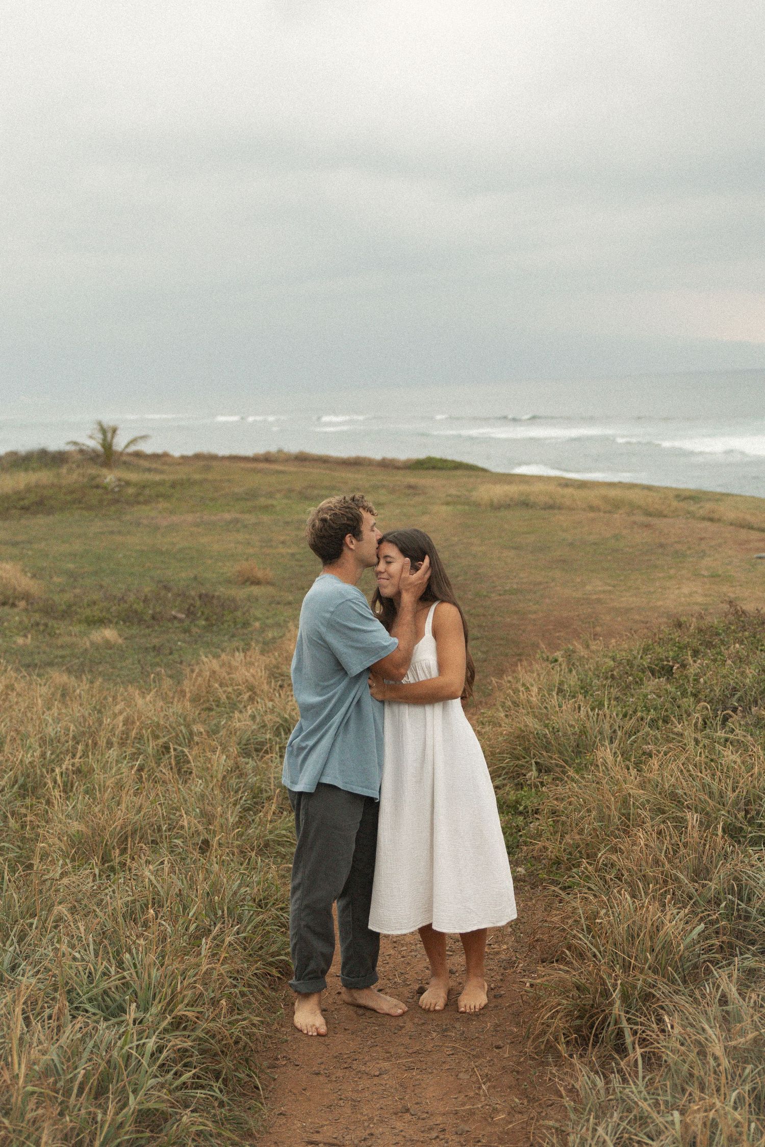 Couple sharing a romantic moment on a coastal path with ocean waves and cloudy skies in the background.