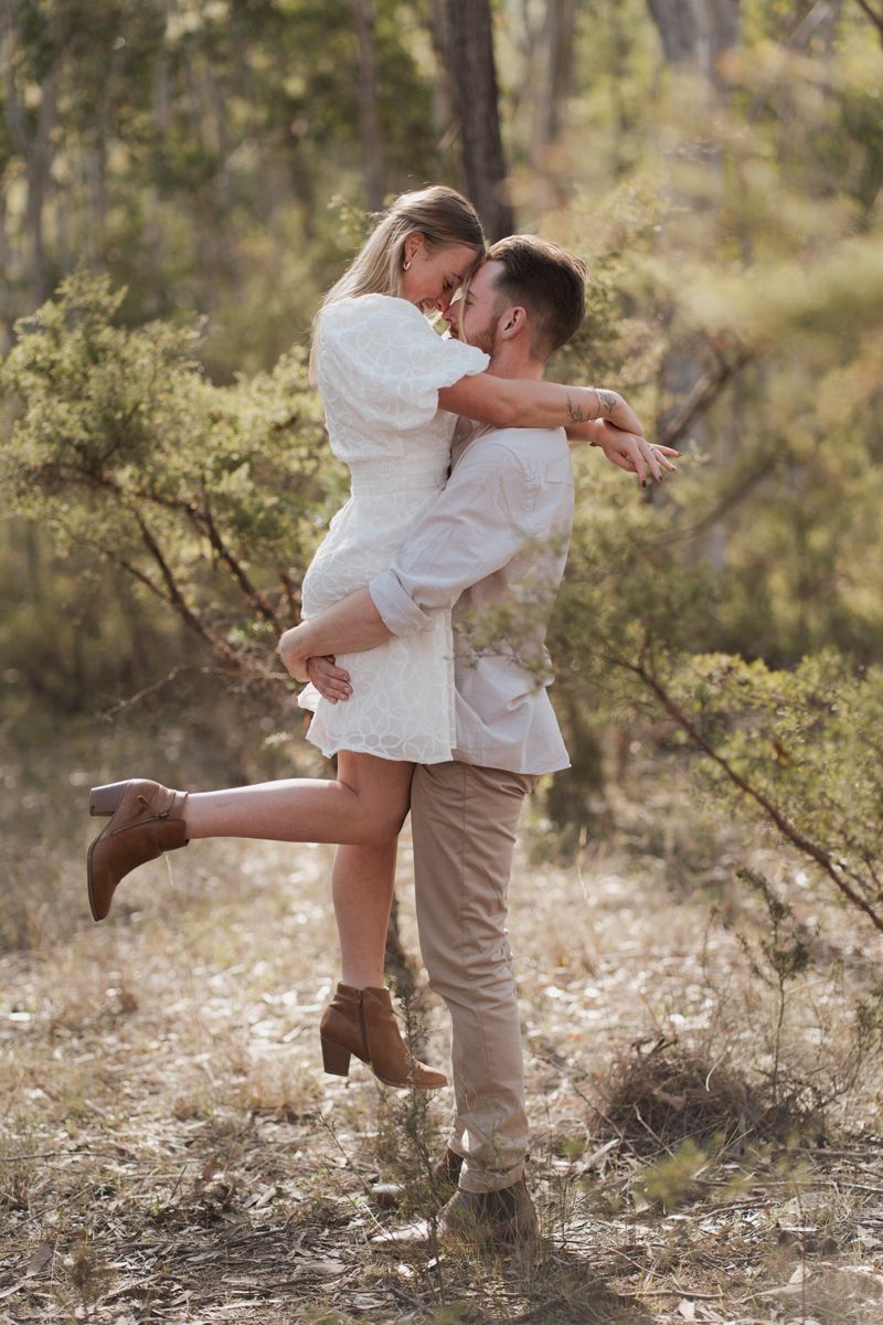 Couple hugging in a sunlit forest, man lifting woman in white dress and brown boots among green trees in rouse hill regional park photoshoot