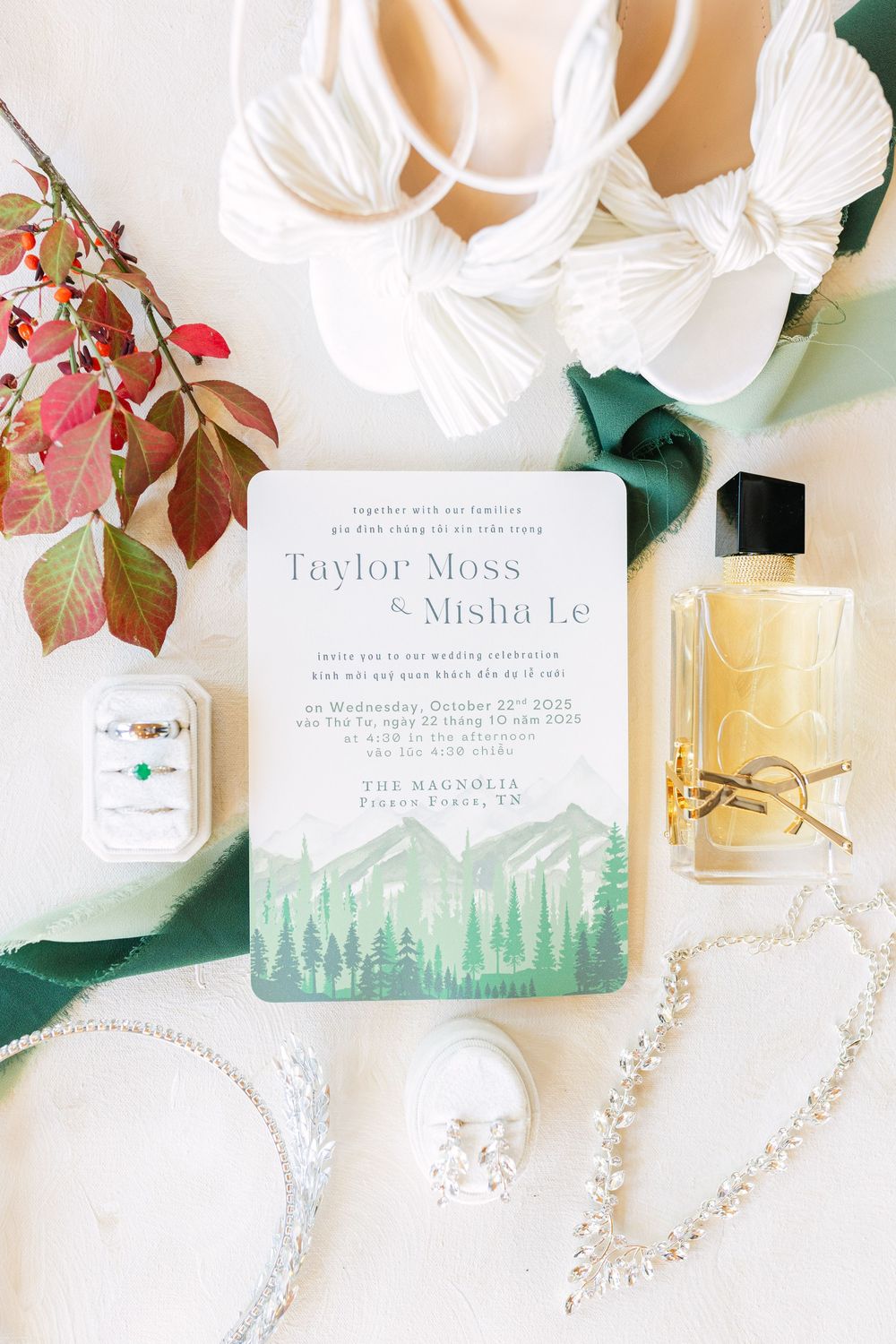 Wedding invitation for Taylor Moss and Misha Le with mountain forest watercolor design, surrounded by rings, perfume, heels, and florals.