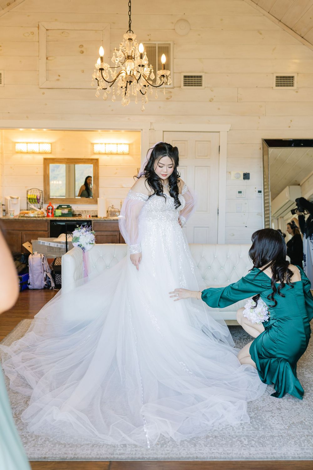 Bride in white ball gown being assisted by bridesmaid in emerald dress inside elegant white wood-paneled bridal suite.