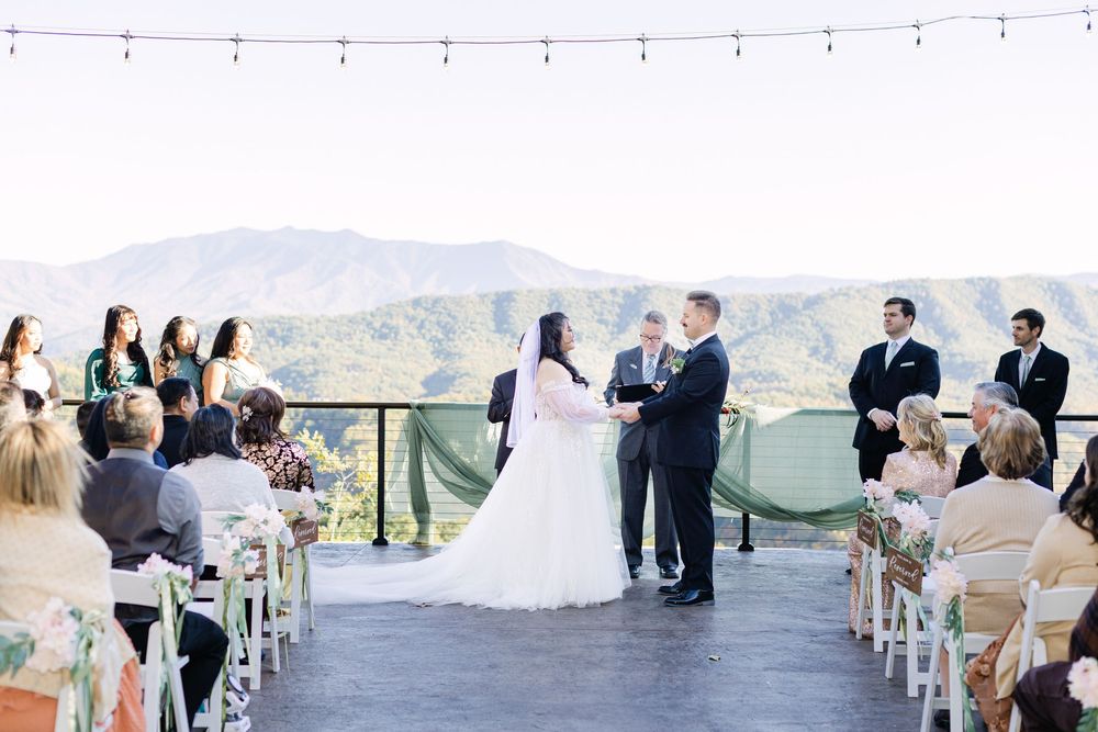 Outdoor wedding ceremony on a scenic mountain overlook with bride and groom exchanging vows, string lights, and guests seated.