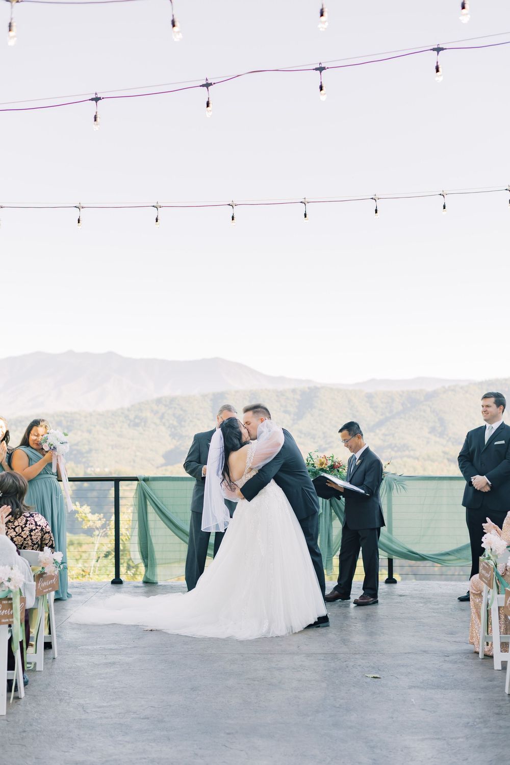 Bride and groom share first kiss at outdoor wedding ceremony with mountain views and string lights.