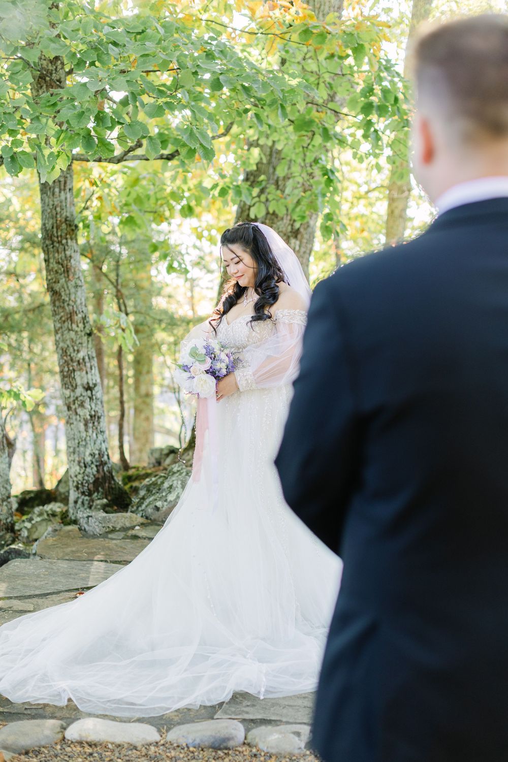 Bride in white gown holding bouquet smiles during outdoor wedding first look among autumn trees.