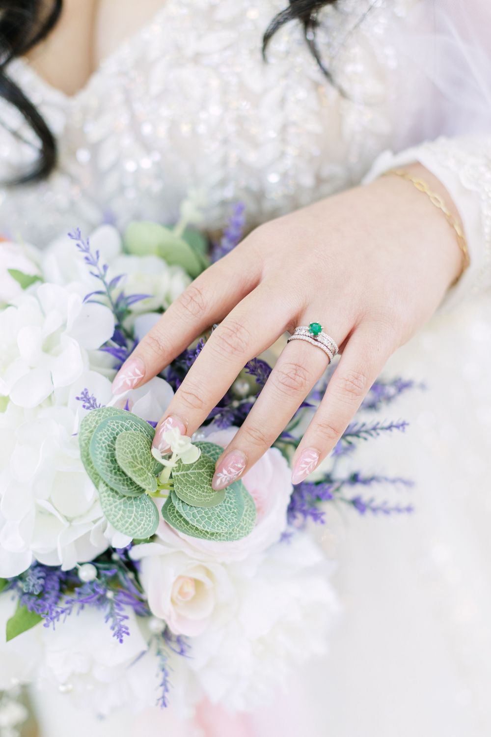 Bride's hand with emerald engagement ring and floral nail art resting on white and purple wedding bouquet.