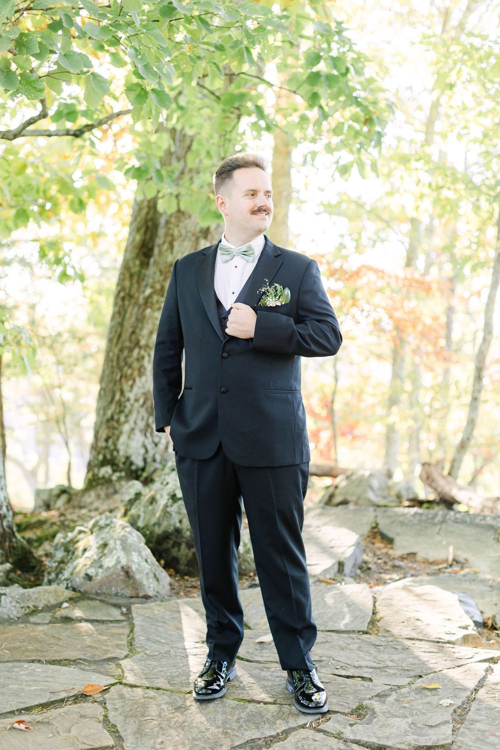 Groom in black tuxedo with mint bow tie and boutonniere standing on stone path surrounded by autumn trees.