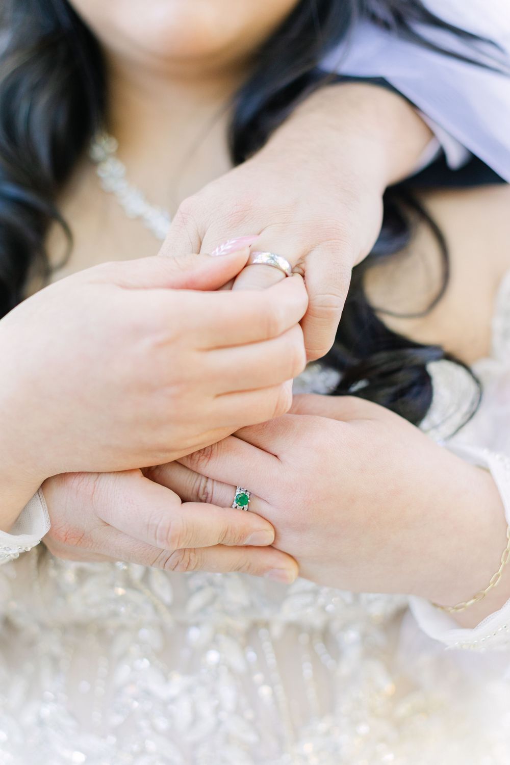 Close-up of bride and groom holding hands showing wedding bands and emerald engagement ring on lace dress.