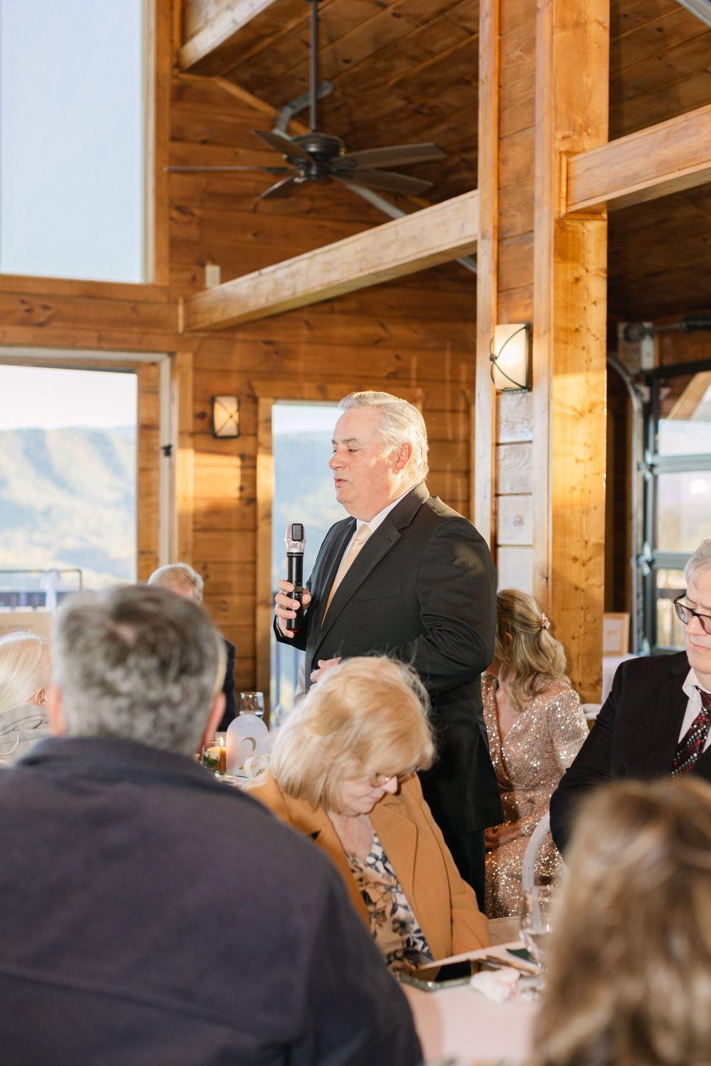 A man in a suit delivers a heartfelt speech with a microphone at a rustic wooden wedding venue.