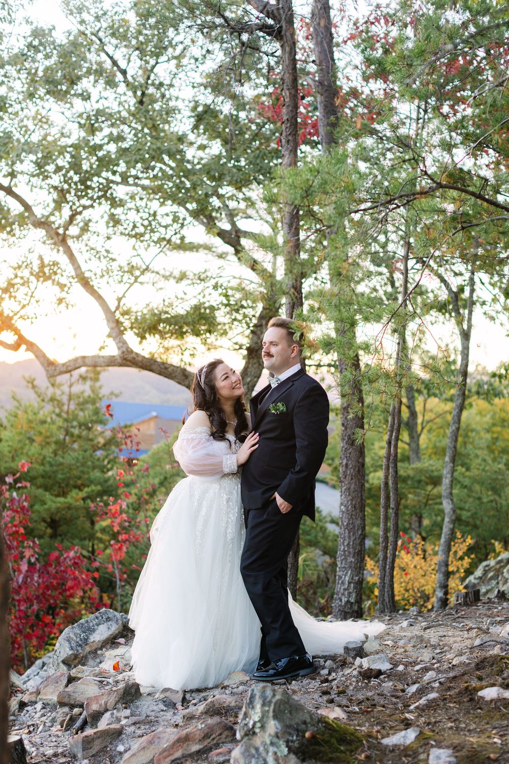 Bride and groom share a romantic moment on a rocky hillside surrounded by colorful autumn trees at golden hour.