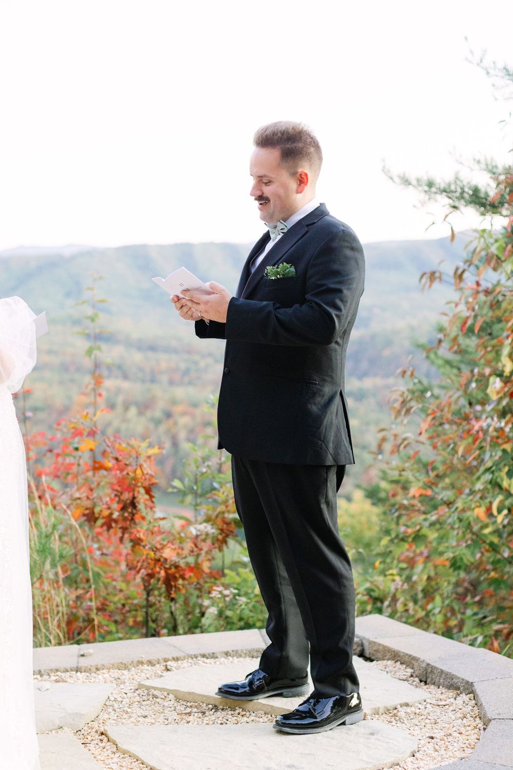 Groom in black suit reads vows outdoors with colorful fall mountain landscape in background.