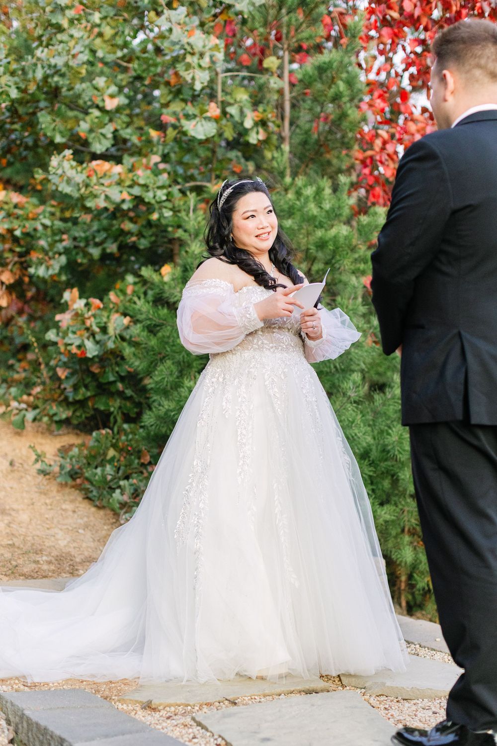 Smiling bride in a beaded ballgown reads vows to her groom amid vibrant fall foliage outdoors.