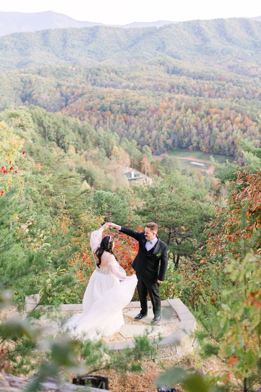 A bride and groom dance together on a stone platform with colorful autumn mountain views behind them.