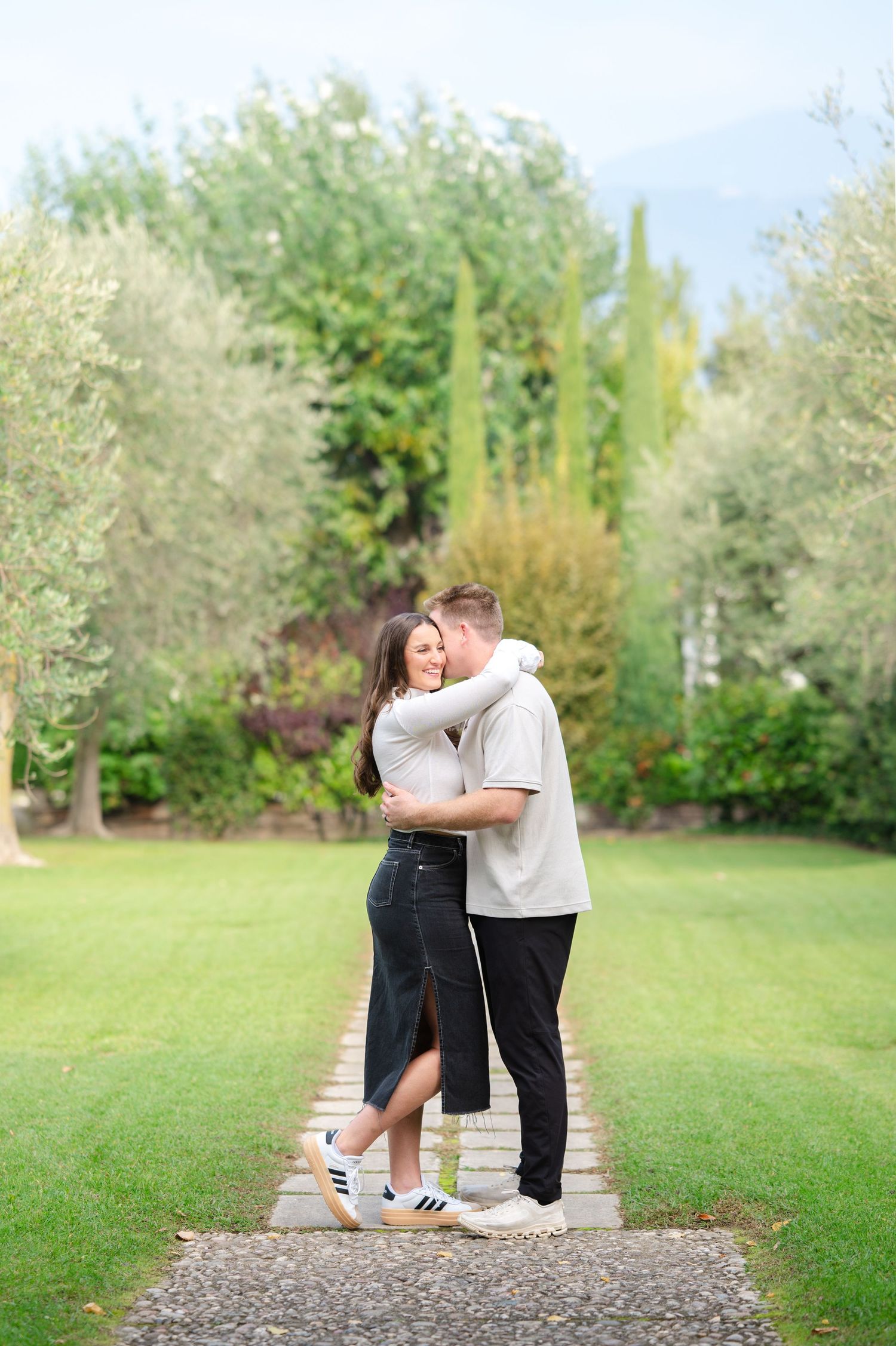A couple embraces and smiles on a garden path surrounded by lush green trees and manicured lawn.