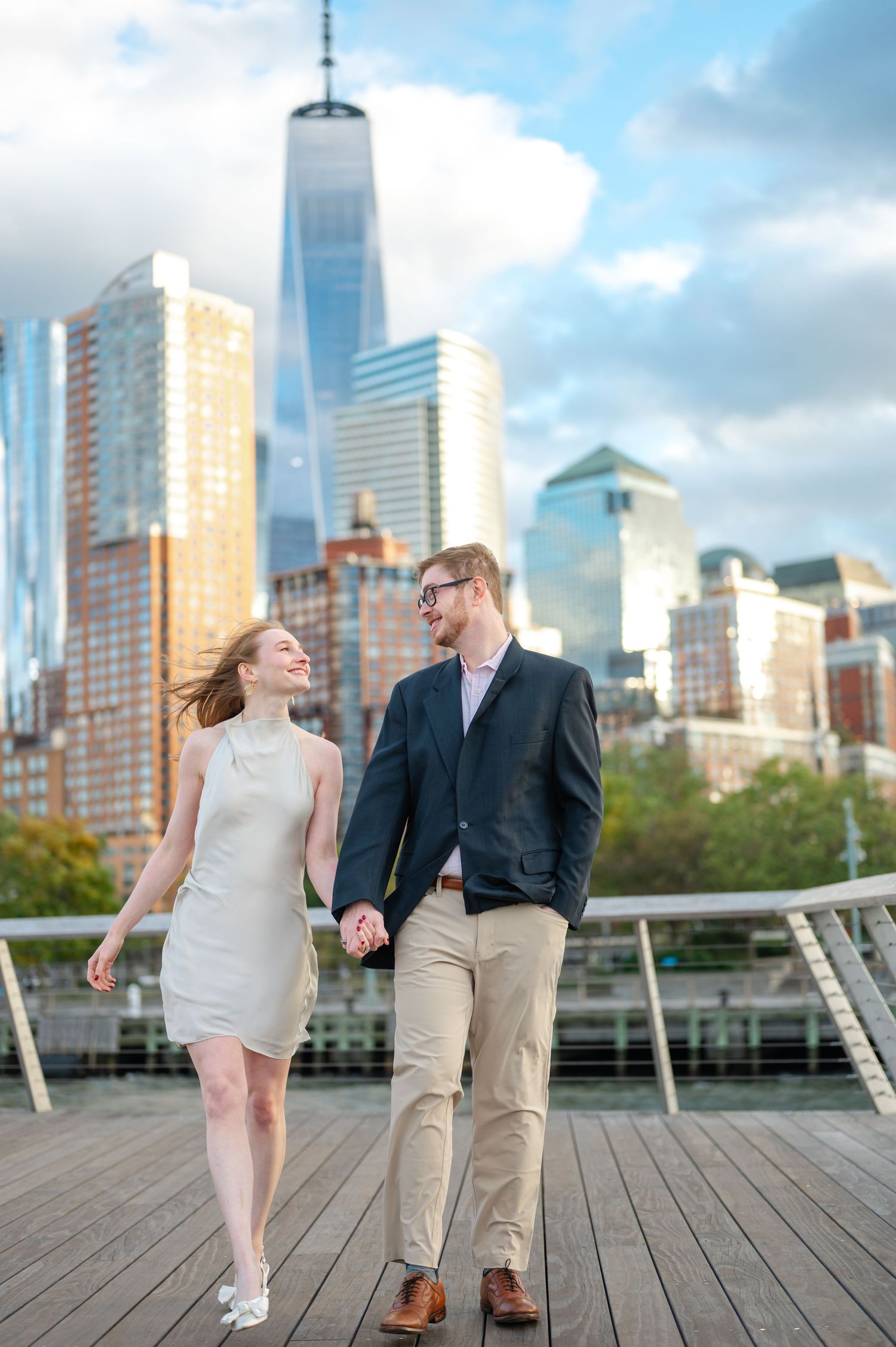 A couple holds hands and walks on a wooden pier with the NYC skyline and One World Trade Center behind them during a NYC engagement session.