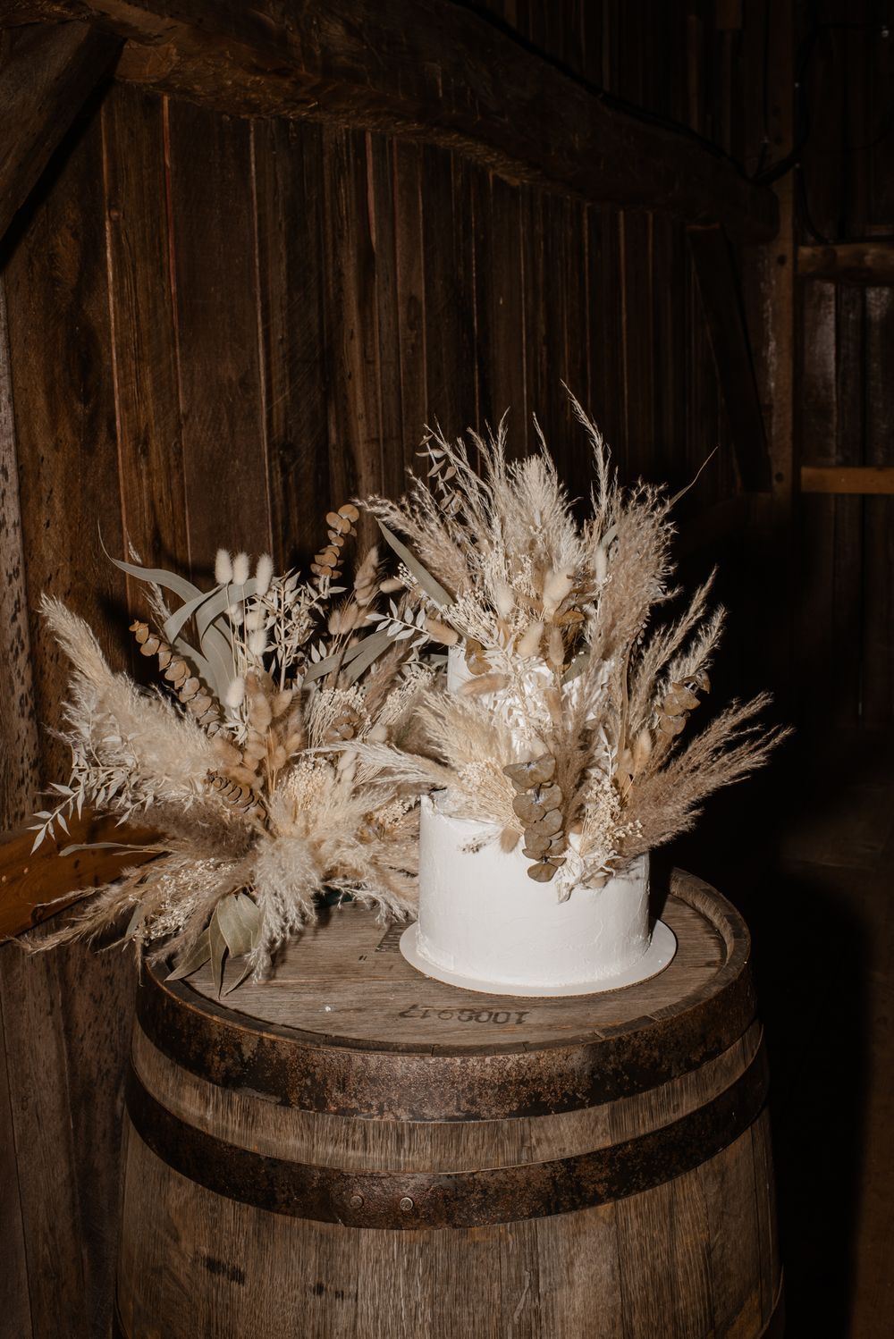 White wedding cake with dried pampas grass on rustic wooden barrel in barn setting.