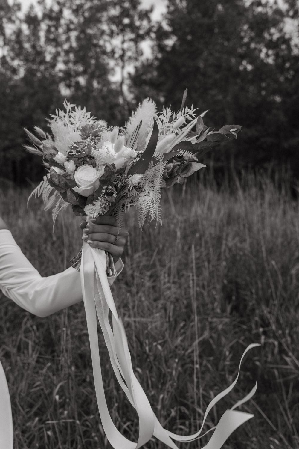 Black and white photo of a bride holding a wildflower wedding bouquet with flowing ribbons in a field.