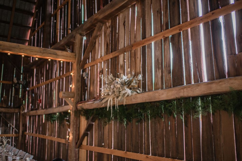Rustic barn wedding venue interior with wooden walls, string lights, greenery garland, and dried floral arrangements on a wooden shelf.