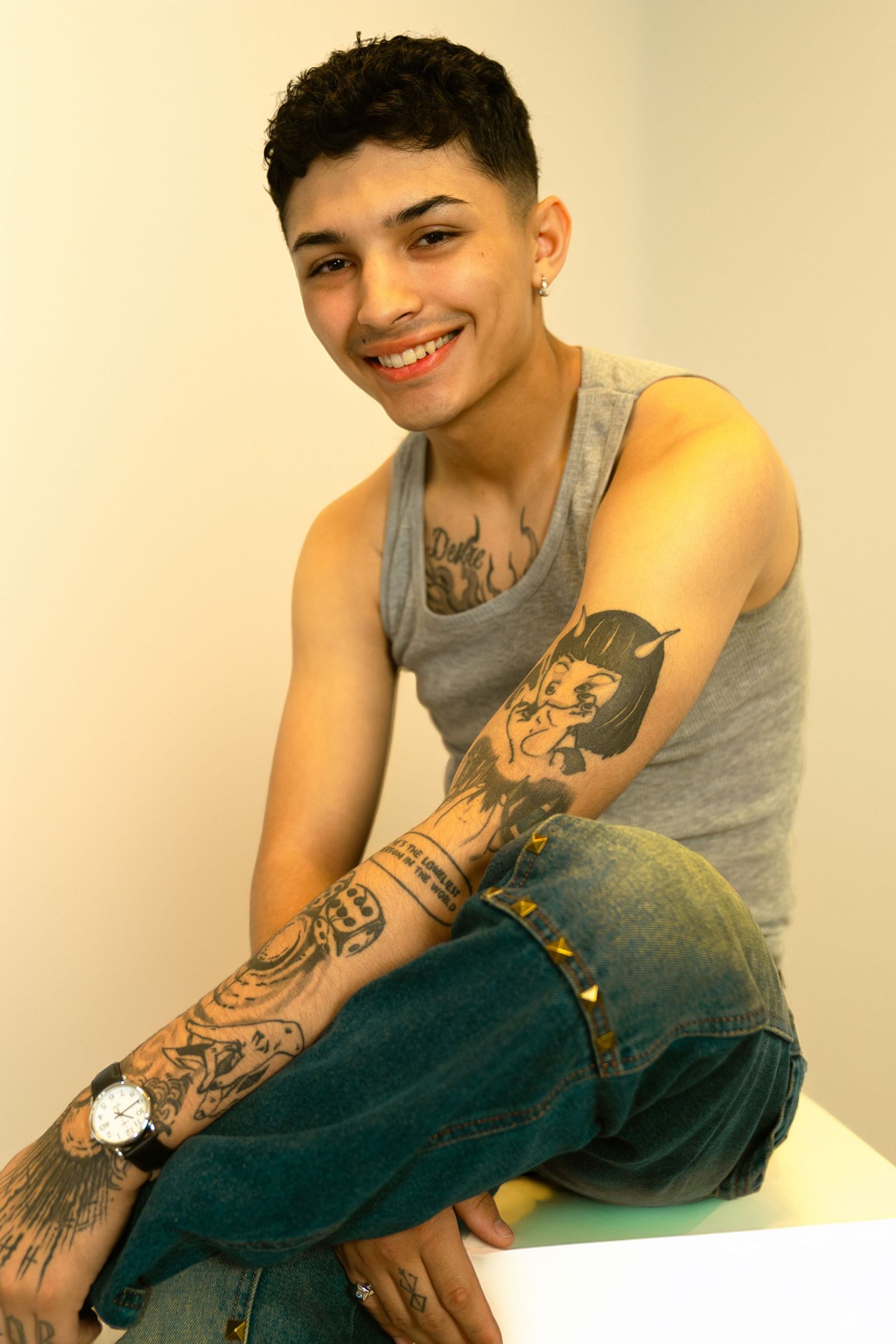 Smiling young man with tattoos on arms wearing gray tank top and jeans, sitting casually while wearing a watch.