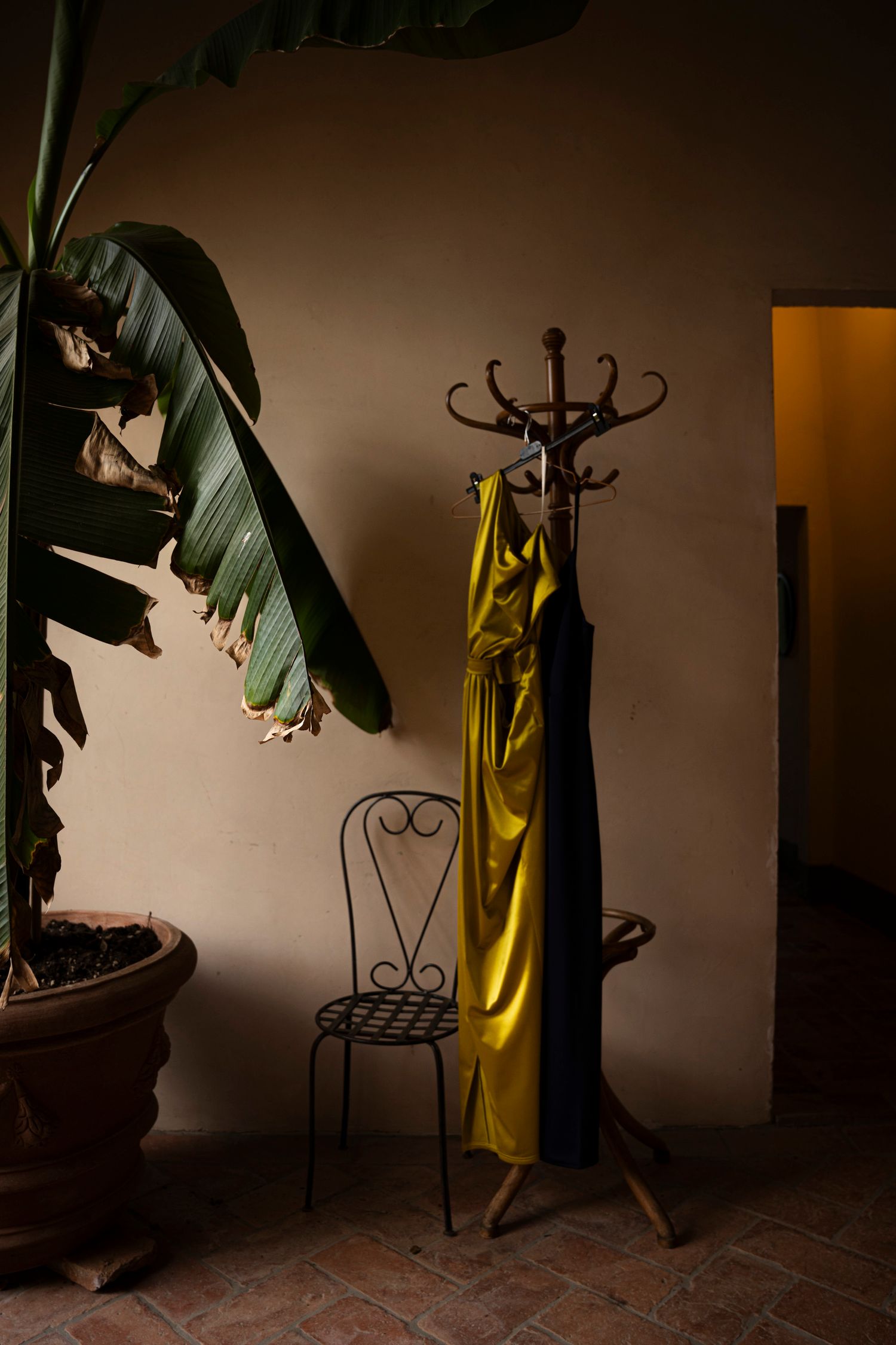 Yellow satin dress draped on vintage coat stand beside tropical plant in dim room.