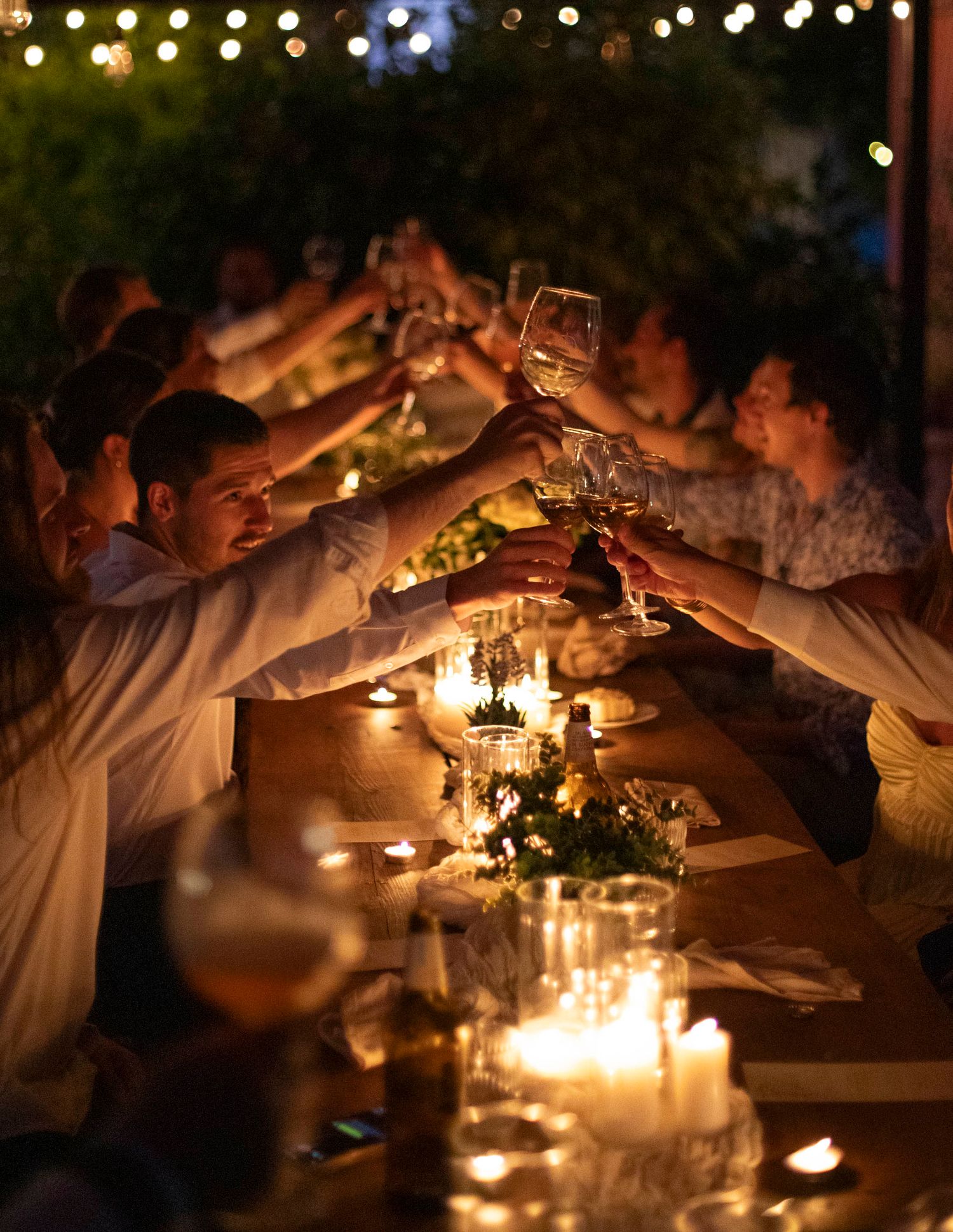 Wedding guests raising glasses in candlelit toast at long outdoor dinner table at night.