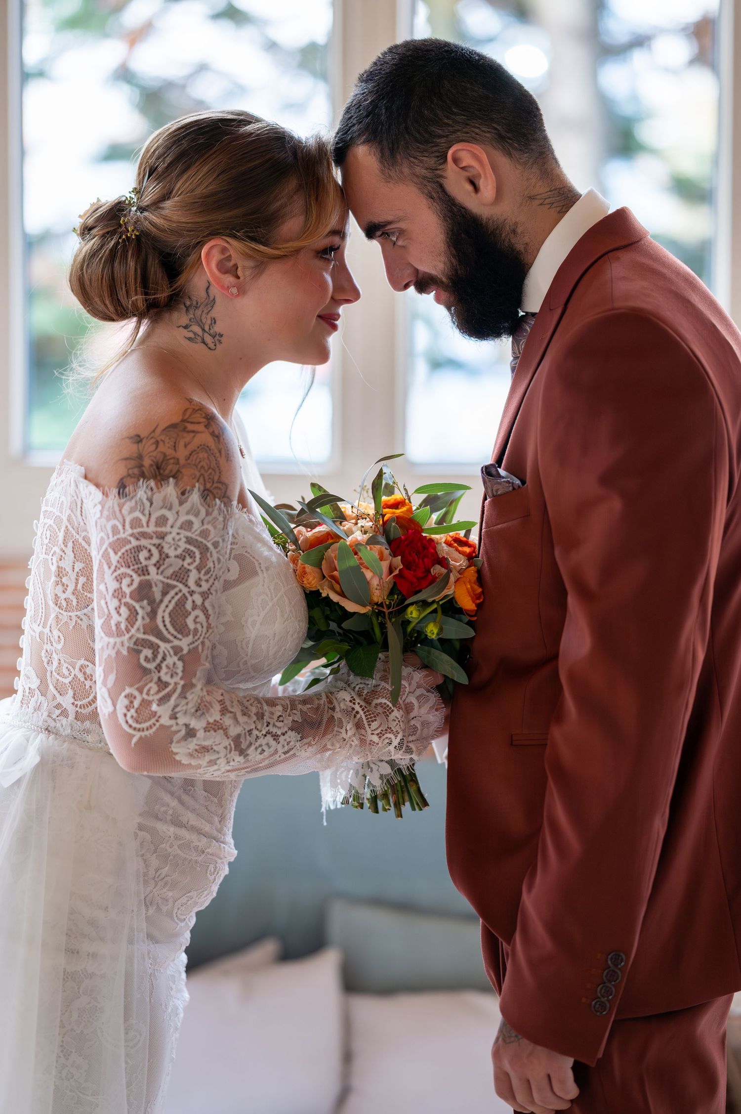 La mariée et le marié touchent tendrement les fronts, elle tient un bouquet coloré, portant une robe en dentelle et un costume rouille.