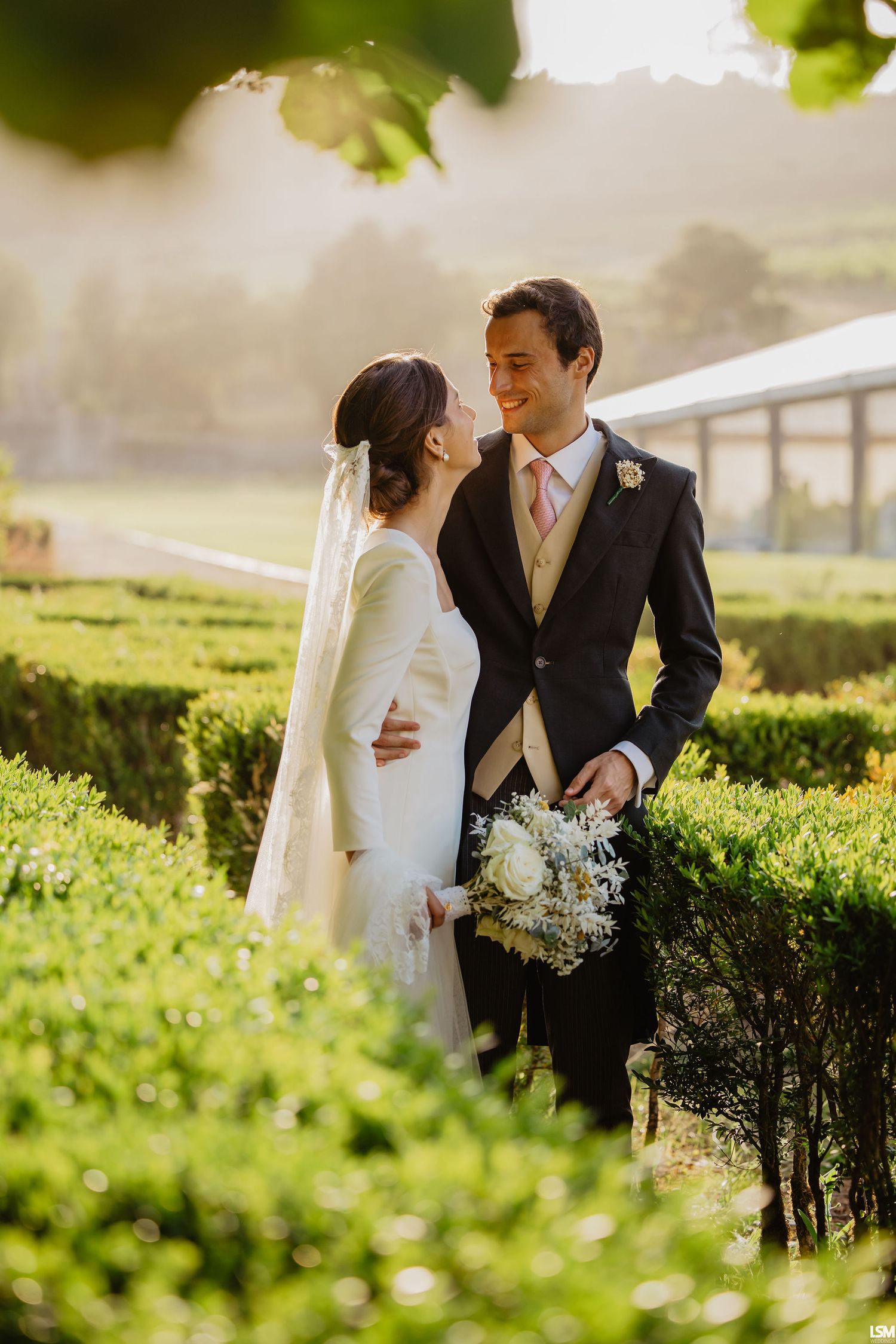 A bride and groom share a romantic moment in a lush garden maze, with soft golden light and a white bouquet.