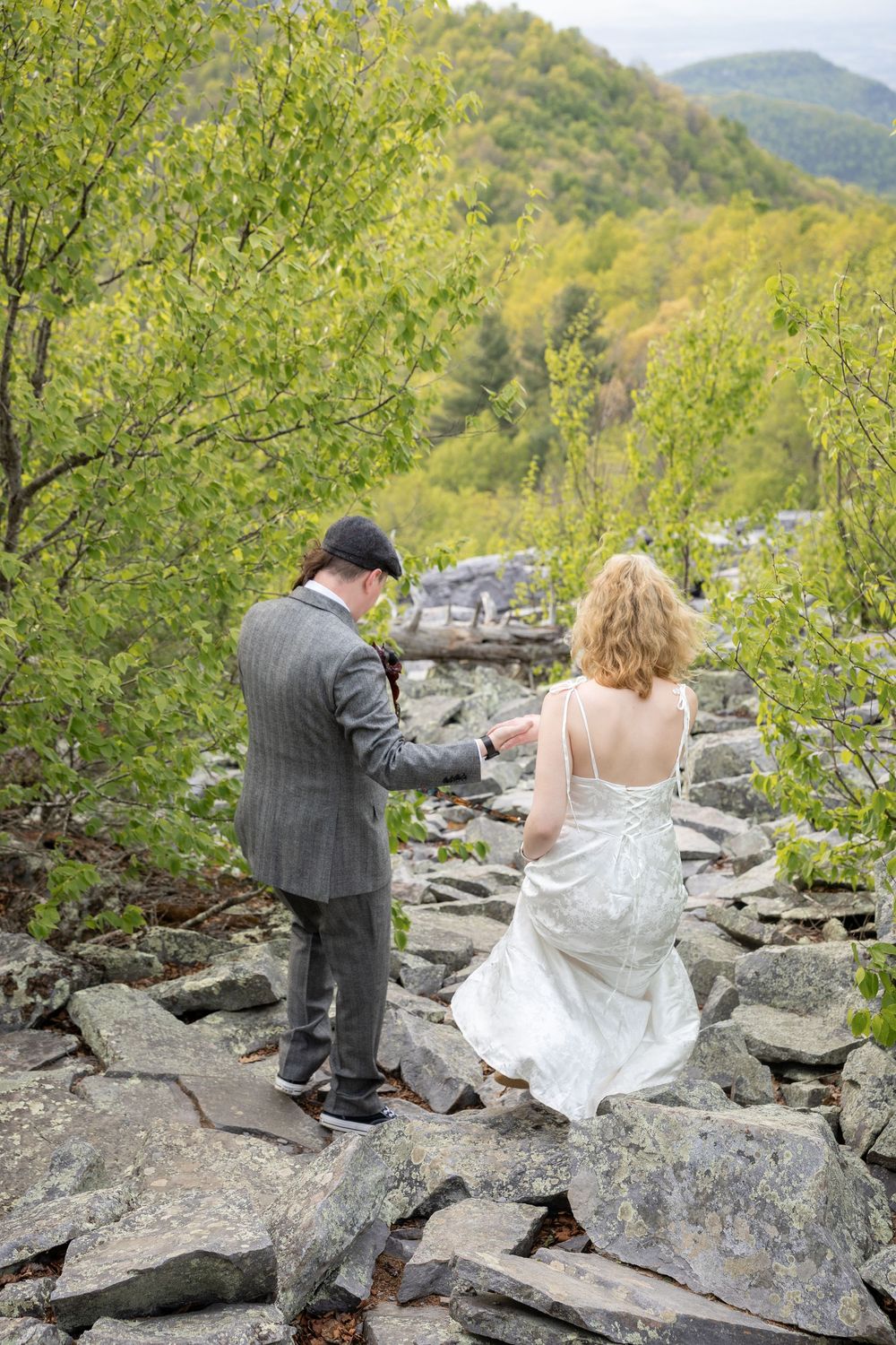 Bride and groom holding hands walking over rocky terrain outdoors, surrounded by lush green trees and mountain scenery.
