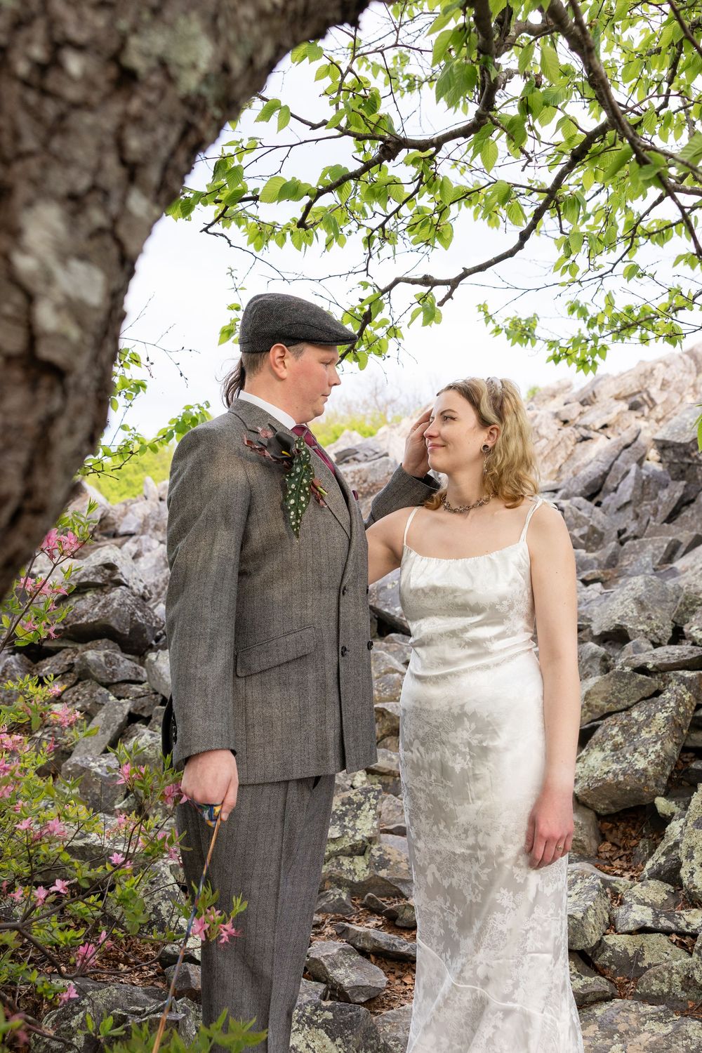 Bride and groom standing together outdoors near a stone wall and tree, wearing elegant wedding attire.