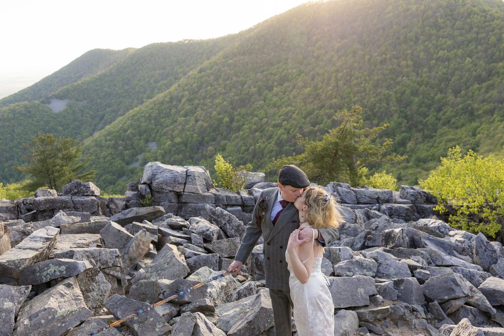 Couple sharing a romantic kiss on rocky mountain summit with lush green hills and golden sunset in background.