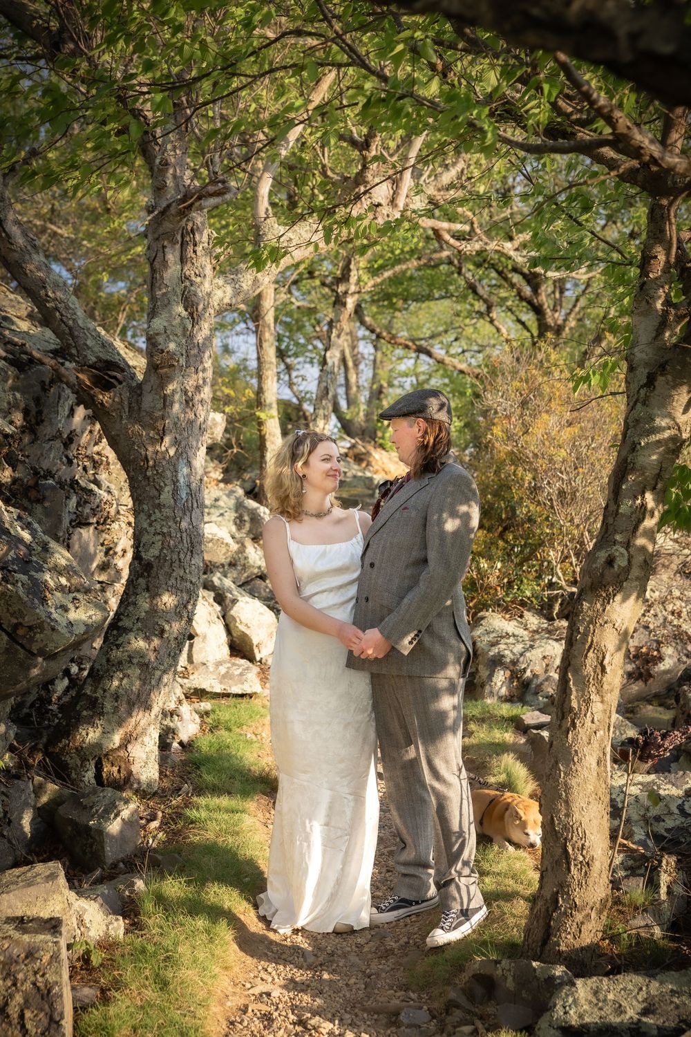 A couple in wedding attire holds hands among rocky woodland trees, the bride in white and groom in a gray suit.