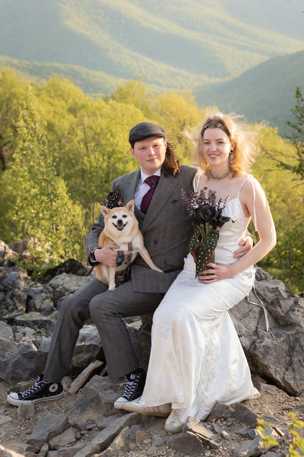 Newlywed couple in wedding attire sitting on rocks with their Shiba Inu dog, mountain forest backdrop.