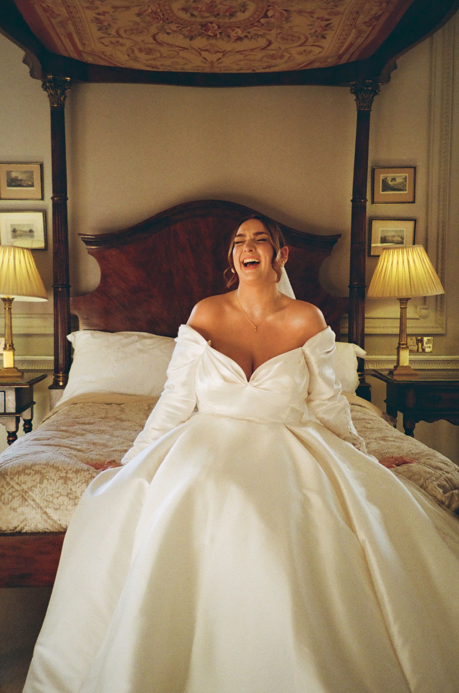 Bride in off-shoulder white ball gown laughing joyfully while sitting on bed in elegant room.