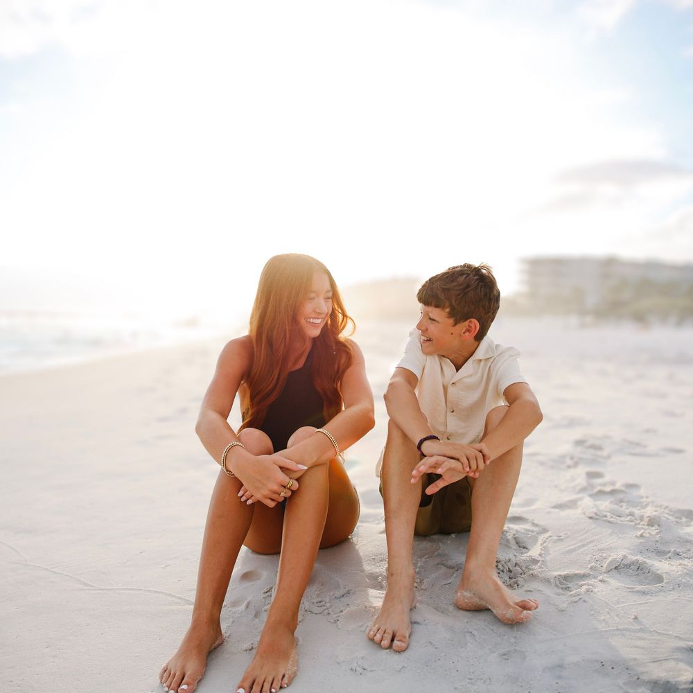 Two children sitting together on a sunny beach, laughing and enjoying a warm golden hour conversation by the shore.