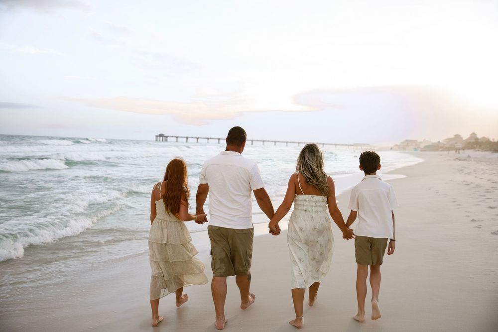 Family of four walking hand-in-hand along a sandy beach at sunset near a pier.