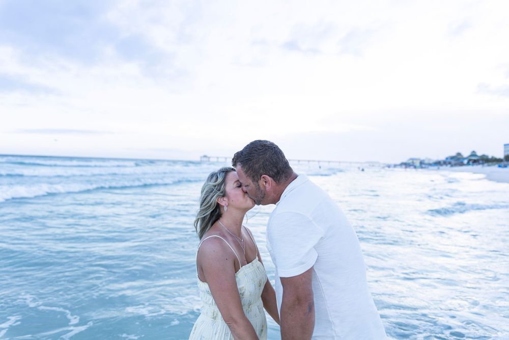 Couple sharing a romantic kiss on a sandy beach at sunset with ocean waves in the background.
