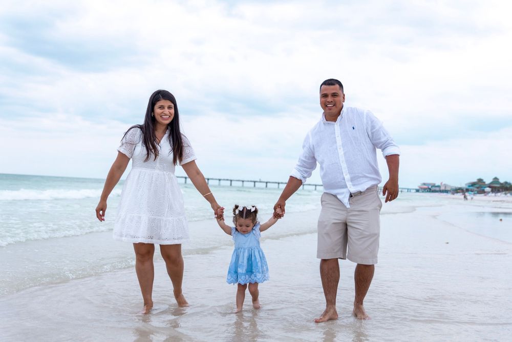 Family of three holding hands on a sandy beach, parents in white outfits with toddler in blue dress, ocean backdrop.